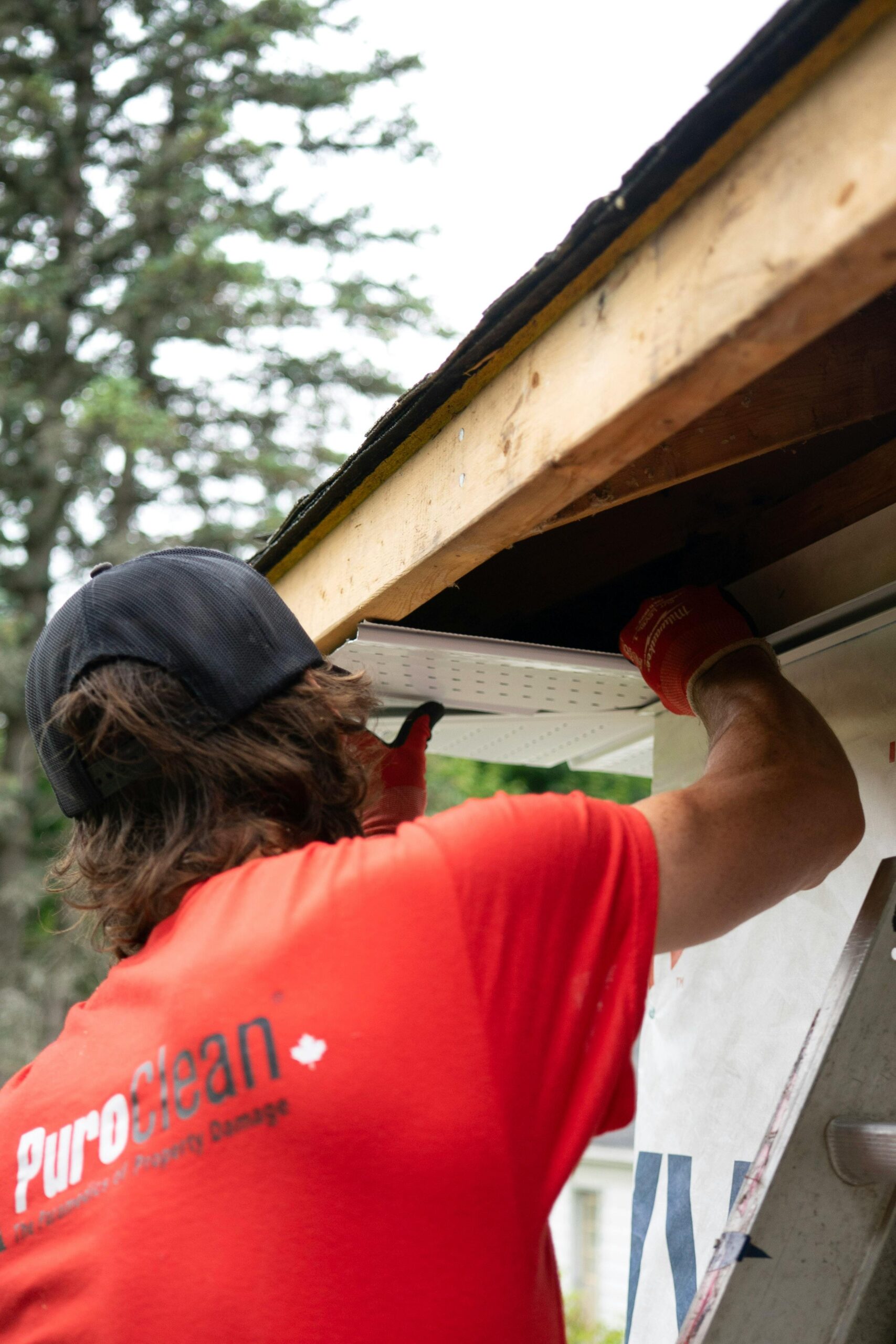 About A construction worker in a red shirt installs a gutter under a wooden roof, focusing on precision.